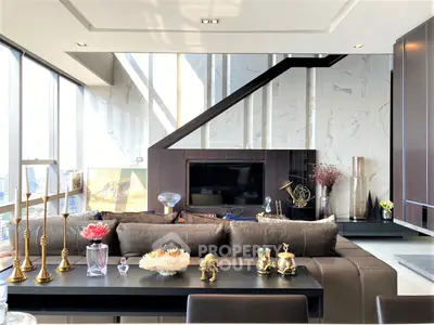 Luxurious living room with modern furniture, elegant decor, and a sleek staircase leading to an upper level, bathed in natural light from floor-to-ceiling windows.