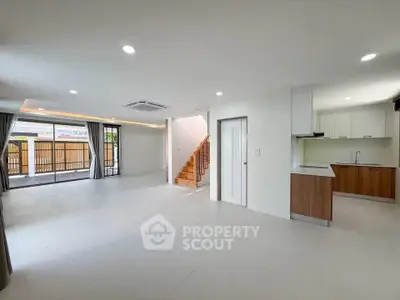 Spacious open-plan living room with modern kitchen and staircase, featuring large windows and ample natural light.