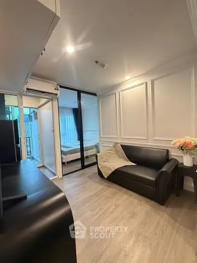 Modern living room with sleek black sofa and elegant panel walls, leading to a cozy bedroom with glass partition.