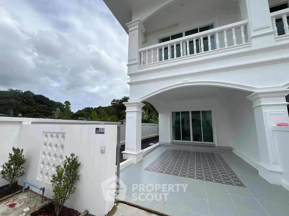 Elegant white two-story house with spacious balcony and tiled patio, surrounded by lush greenery.