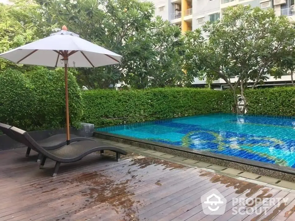 Luxurious outdoor pool area with sun loungers and umbrella, surrounded by lush greenery in a modern residential complex.