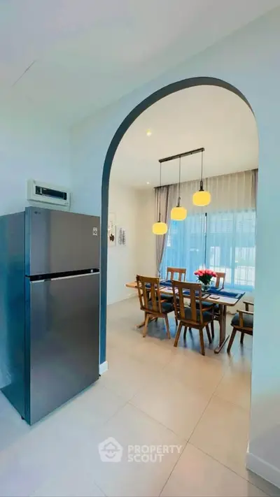 Modern dining area with elegant lighting and large windows, adjacent to a sleek kitchen with stainless steel fridge.