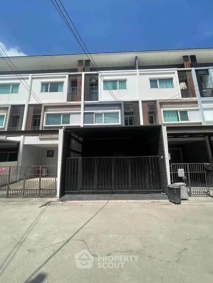 Modern multi-story townhouse with sleek facade and gated parking area under clear blue sky.