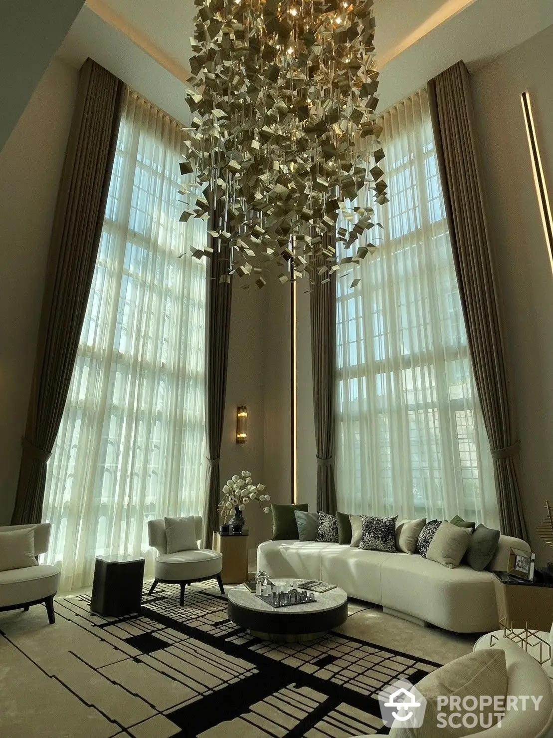 Luxurious living room with high ceilings, grand chandelier, floor-to-ceiling windows draped in sheer curtains, and elegant modern furniture.