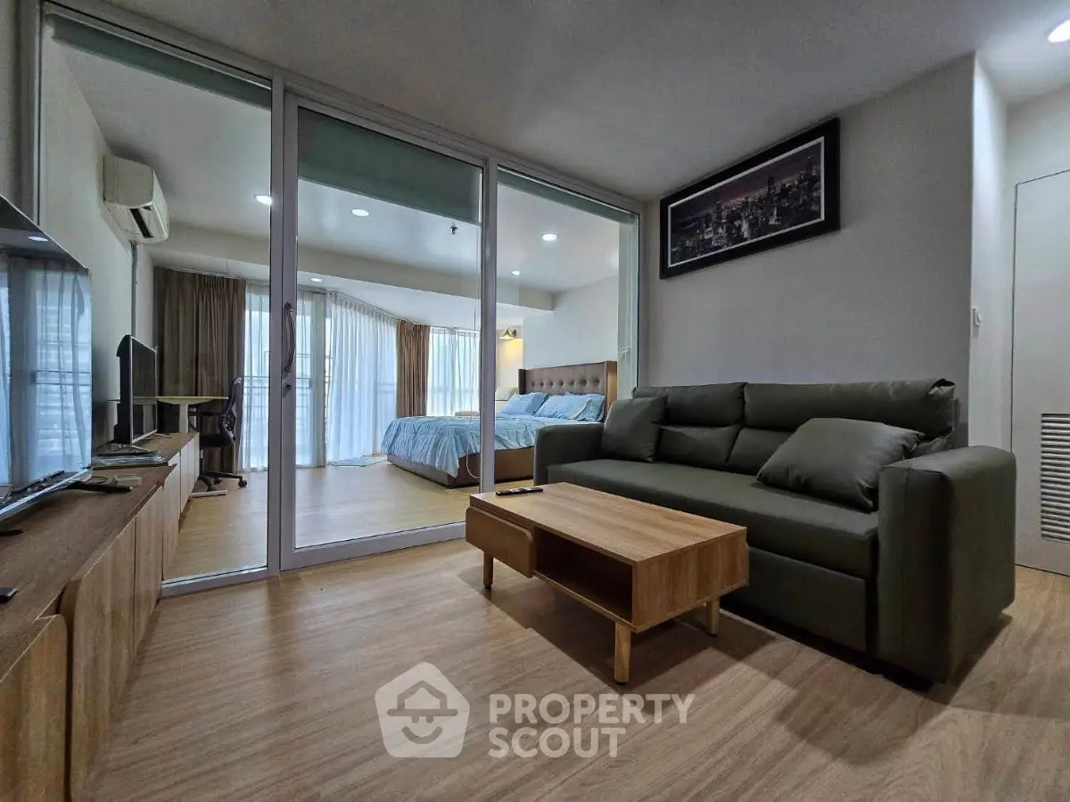 Modern living room with glass partition leading to a cozy bedroom, featuring stylish furniture and wooden flooring.