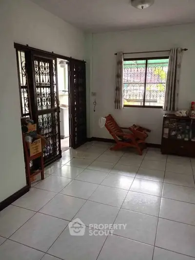 Spacious living room with tiled floor and large window, featuring wooden chairs and a glass cabinet.