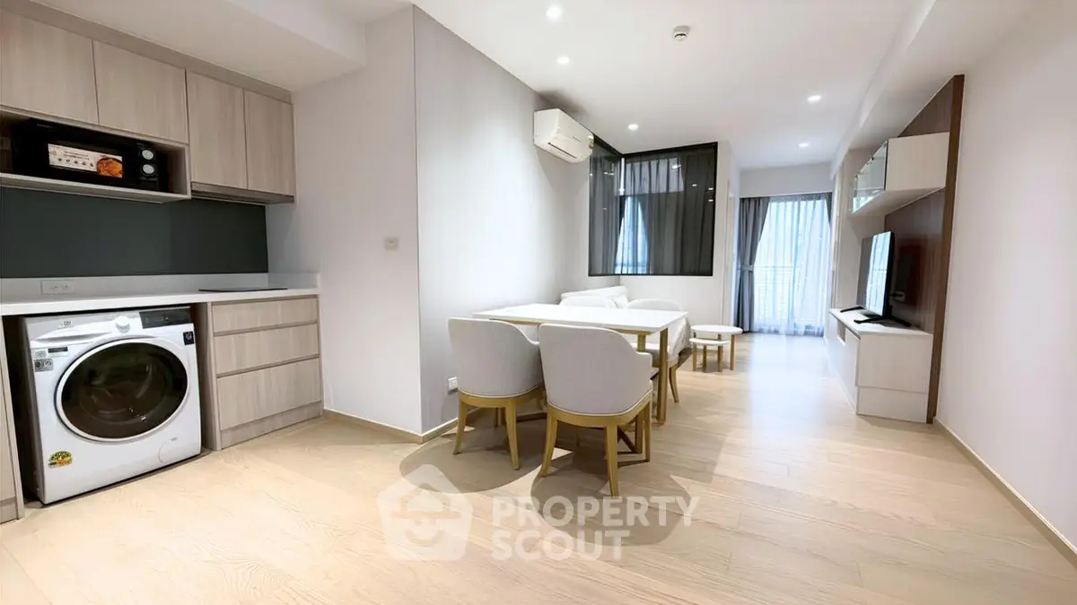 Modern living room with open kitchen and washing machine, featuring sleek furniture and bright lighting.