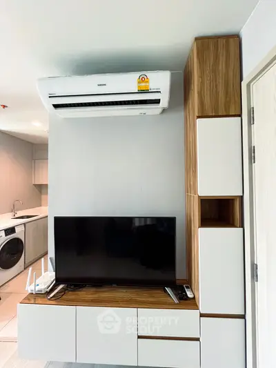 Modern living room with sleek TV unit and air conditioning, adjacent to a compact kitchen with washing machine.