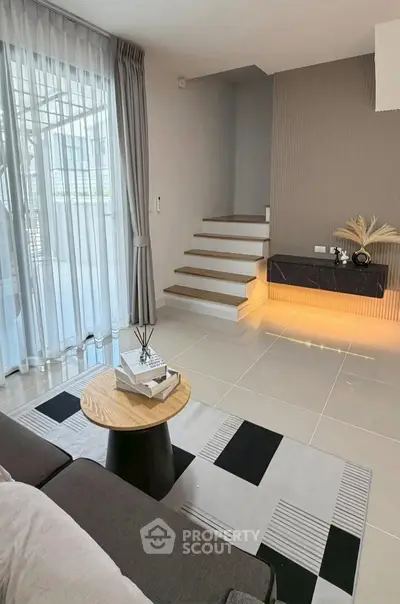 Modern living room with stylish decor and staircase, featuring large windows and elegant curtains.