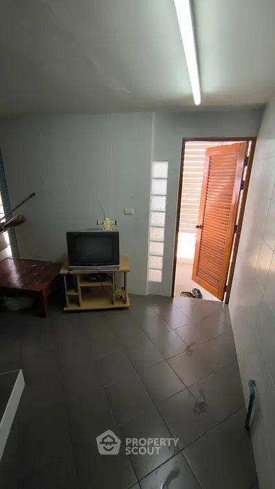 Compact living space with TV and open door leading to a bright area, tiled flooring and minimal furnishings.