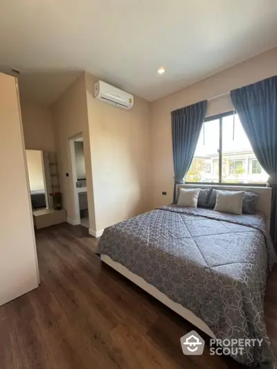 Spacious bedroom with polished wooden flooring, large windows with elegant drapes, and a modern air conditioning unit, offering a serene and comfortable living space.