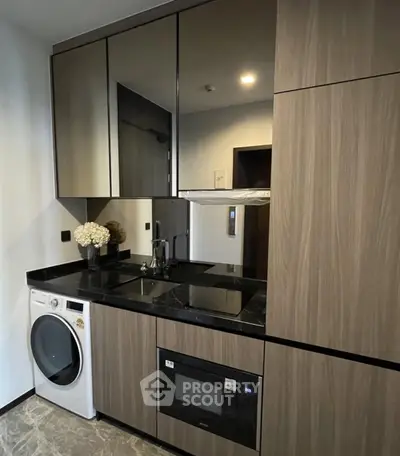 Modern kitchen with sleek appliances and wood cabinetry, featuring a washing machine and microwave.