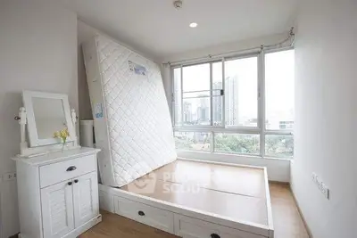 Bright bedroom with large windows and city view, featuring a white dresser and a bed frame without mattress.
