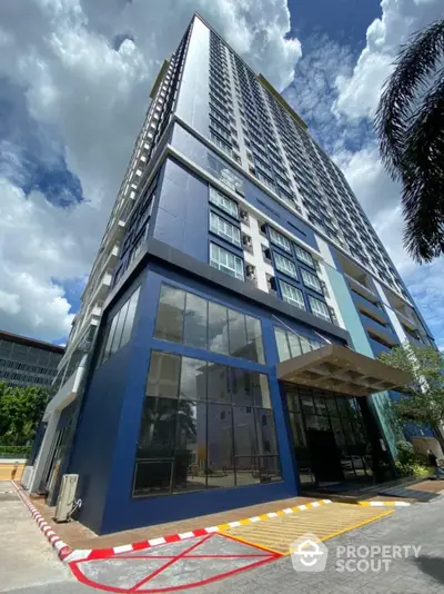 Modern high-rise condominium with a striking blue facade under a sunny sky, showcasing urban living with a stylish entrance and lush greenery.