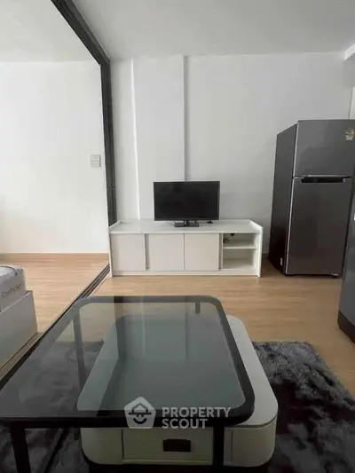 Modern living room with sleek glass coffee table and compact TV setup, adjacent to a stainless steel fridge.