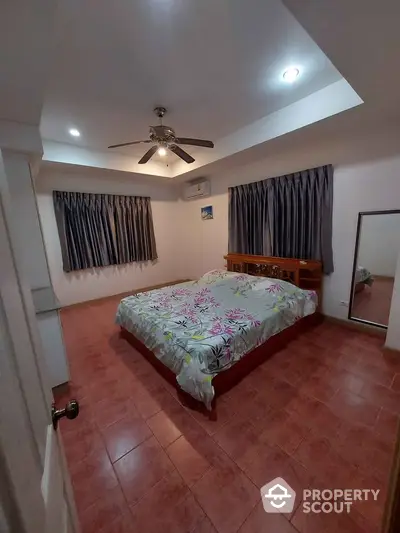 Spacious bedroom with terracotta tiled flooring, a large bed with patterned bedding, ceiling fan, and ample natural light from curtained windows.