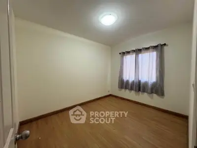 Spacious empty bedroom with wooden flooring and natural light through curtained window.