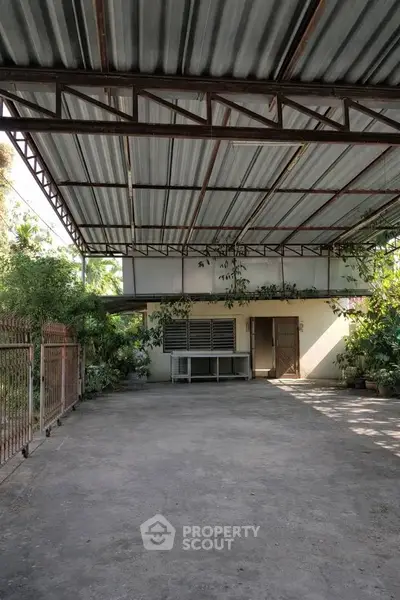 Spacious covered outdoor area with metal roof and lush greenery, ideal for parking or gatherings.