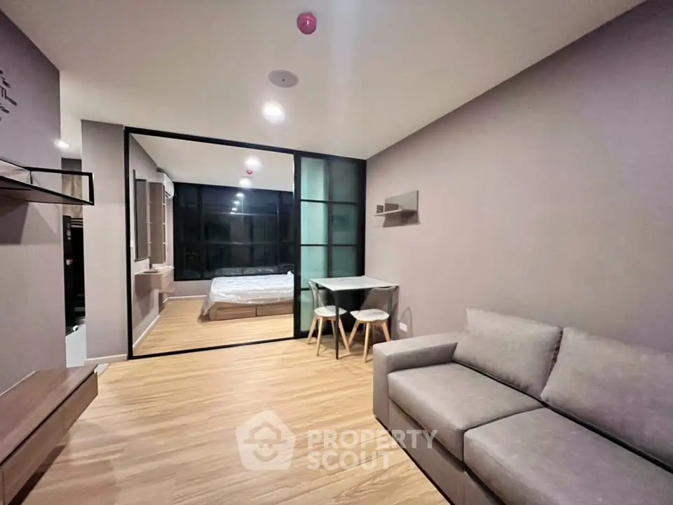 Modern apartment interior with cozy living room and glass-walled bedroom, featuring stylish furniture and wooden flooring.