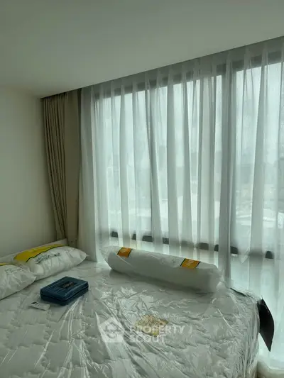 Cozy bedroom with large window and sheer curtains offering natural light and city view.