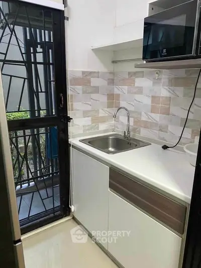 Compact modern kitchen with sleek cabinetry and tiled backsplash, featuring a built-in microwave and stainless steel sink.