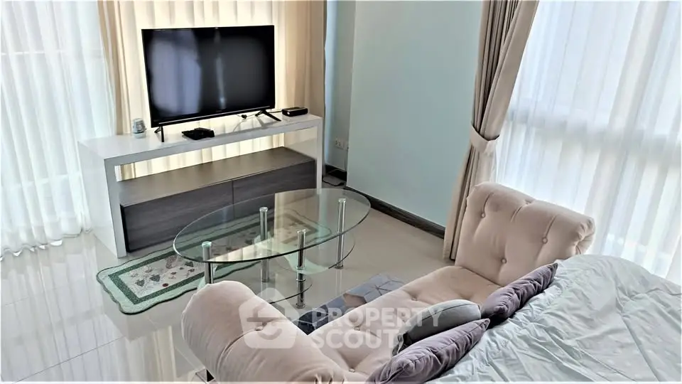 Modern living room with plush sofa, glass coffee table, and large TV, bathed in natural light from floor-to-ceiling windows.