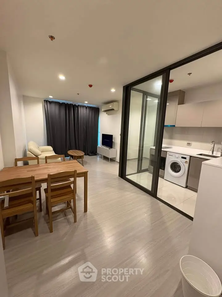 Modern apartment interior with open living and dining area, featuring a compact kitchen and washing machine.