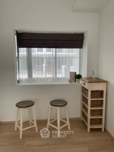 Bright and airy minimalist kitchen space with natural light streaming through a large window, complemented by modern wooden stools and storage unit.