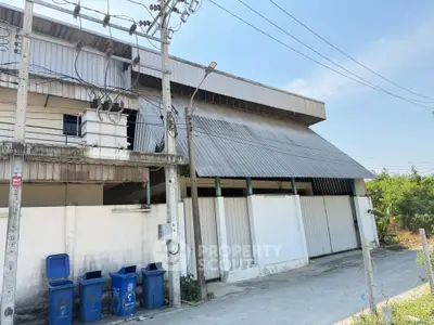 Industrial warehouse exterior with large metal roof and blue recycling bins, ideal for storage or manufacturing.
