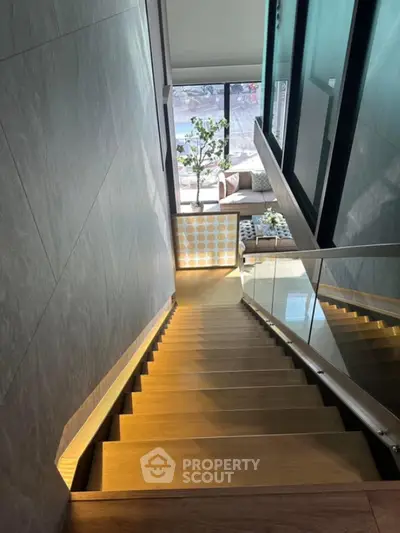 Modern staircase with glass railing leading to a stylish living area with large windows.