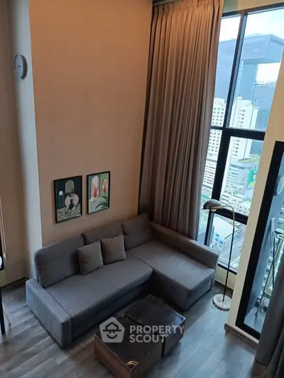 Modern living room with high ceiling and city view, featuring a stylish gray sofa and floor-to-ceiling windows.