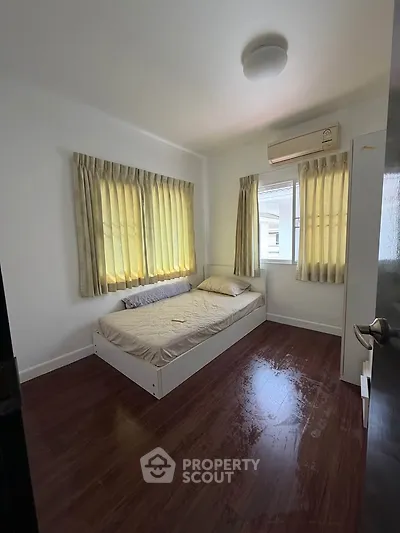 Cozy bedroom with wooden flooring and natural light from large windows, featuring a comfortable bed and air conditioning.
