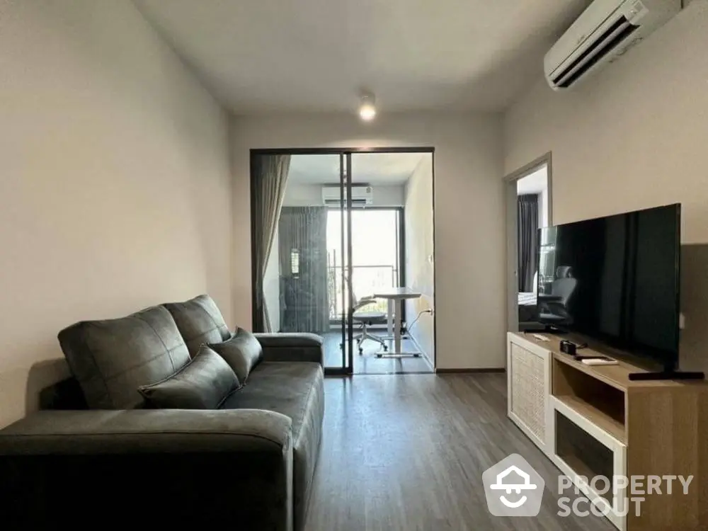 Modern living room with sleek furniture and large TV, leading to a balcony with natural light.