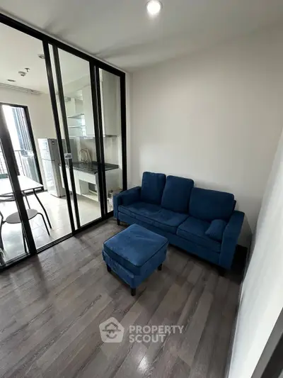 Modern living room with blue sofa and glass partition, leading to a bright kitchen area.