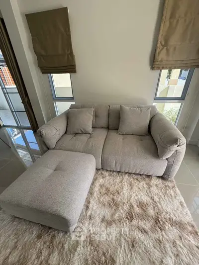 Cozy living room with plush sofa and ottoman on a soft rug, featuring large windows for natural light.