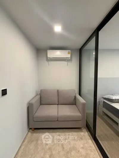 Cozy small living area with modern gray sofa and air conditioning, adjacent to a bedroom with glass partition.