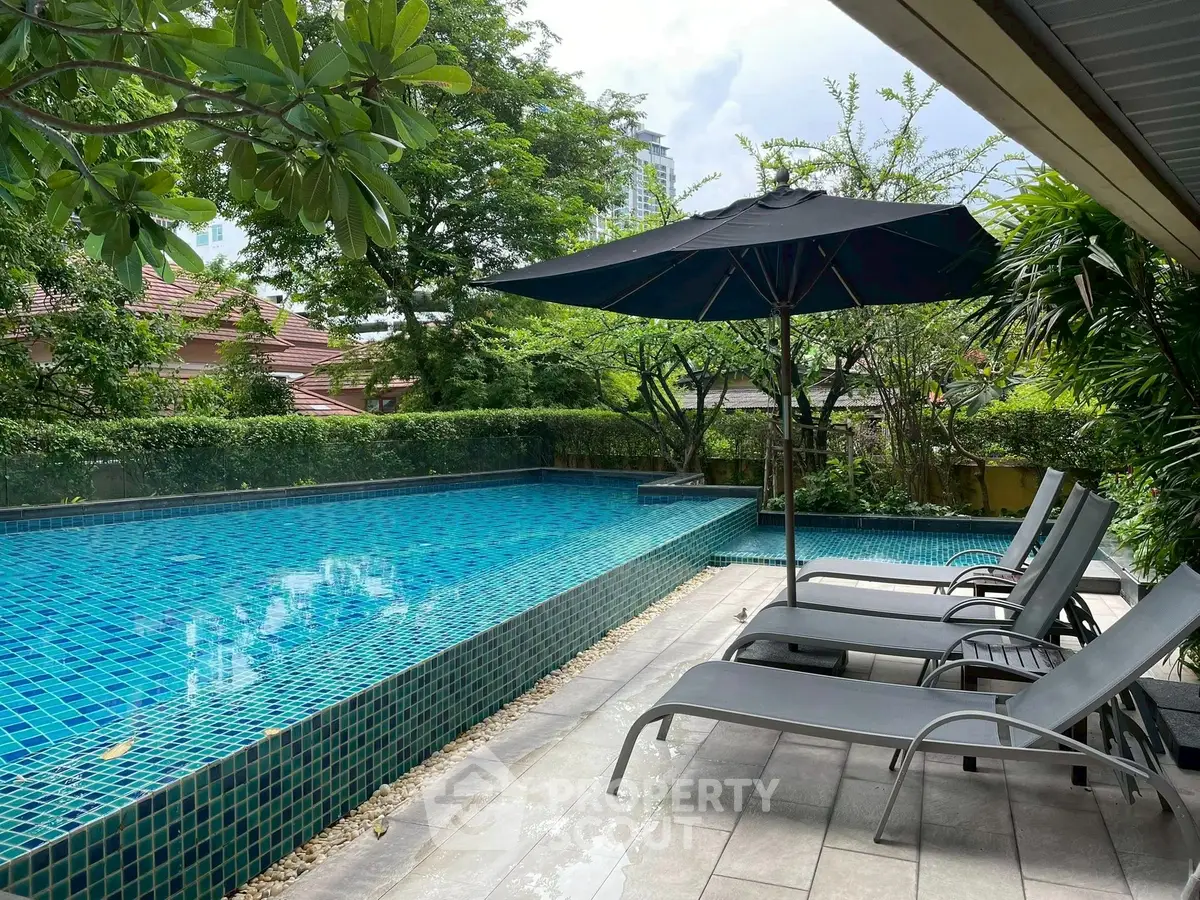 Luxurious outdoor pool area with sun loungers and umbrella, surrounded by lush greenery.