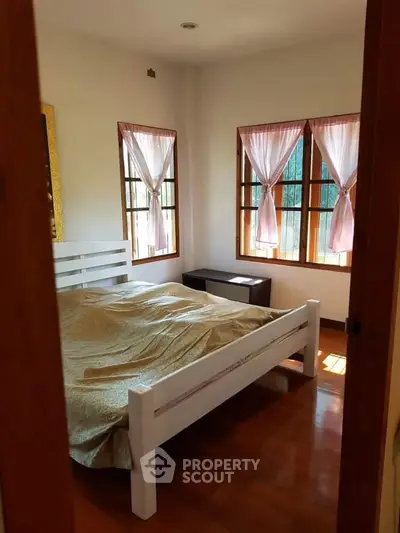 Cozy bedroom with wooden flooring and natural light through curtained windows.