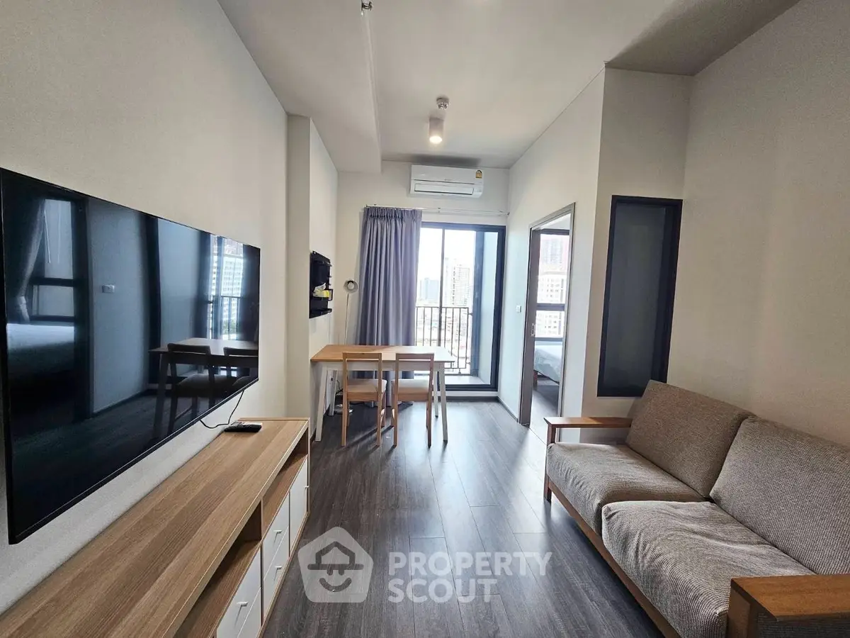 Modern living room with sleek furniture and large TV, leading to a balcony with city views.