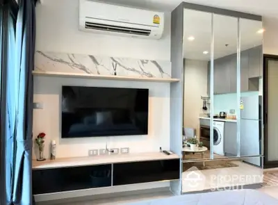 Modern apartment interior with sleek finishes, featuring a mounted TV, mirrored wall, and a glimpse into a well-equipped kitchen with washing machine.