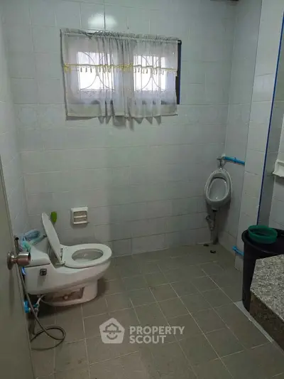 Spacious bathroom with toilet and urinal, tiled walls and floor, small window with curtain.