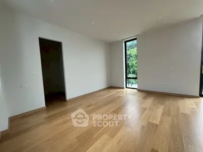 Spacious empty room with wooden flooring and large window offering lush green view.