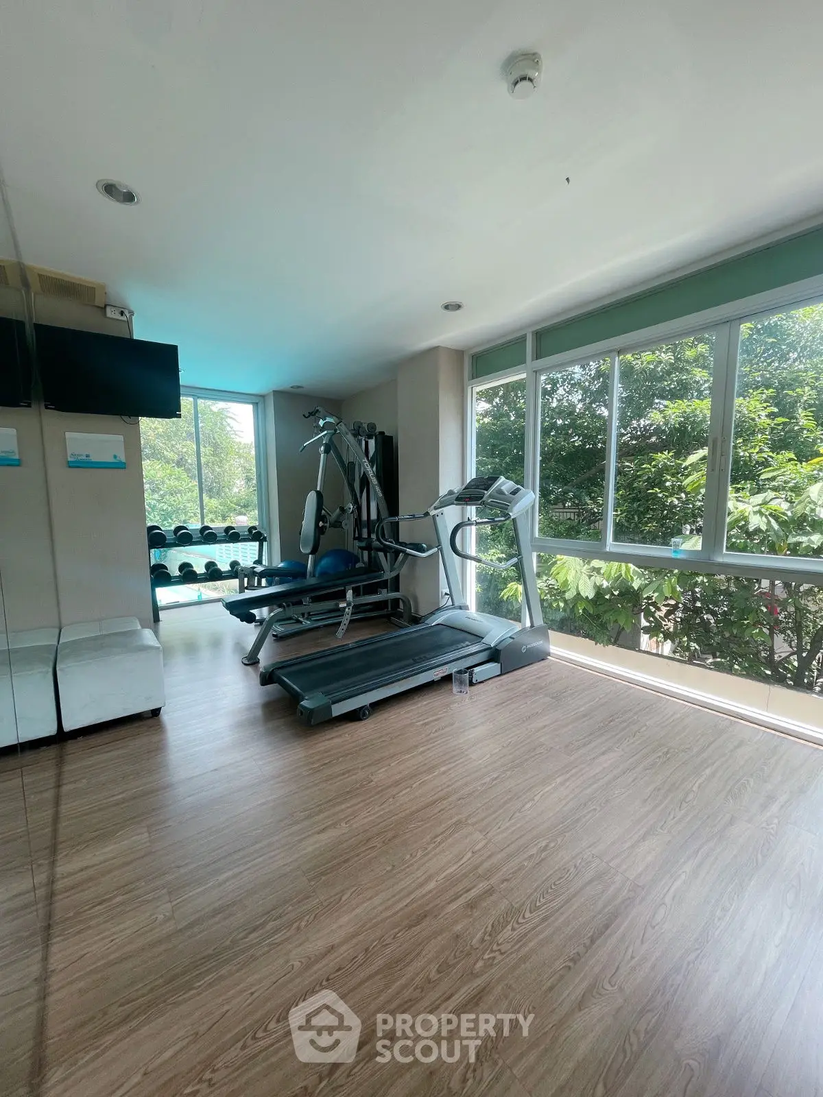 Modern gym with treadmill and weights, surrounded by large windows with lush greenery view.