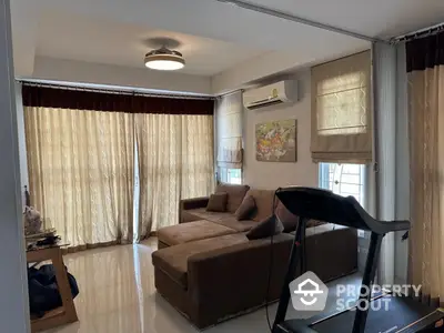 Spacious living room with modern sofa and treadmill, featuring large windows and elegant curtains.