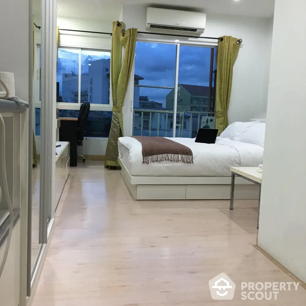 Modern bedroom with large window and city view, featuring cozy bed and workspace.