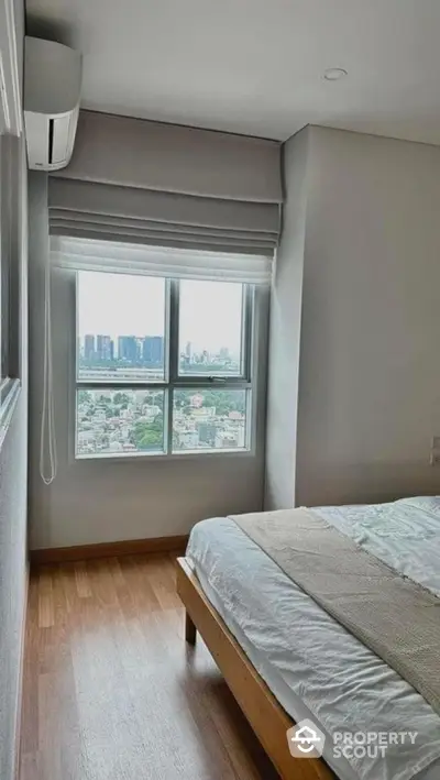 Cozy bedroom with city view from large window, featuring wooden flooring and modern air conditioning.