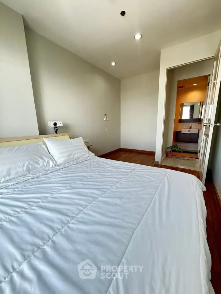 Cozy bedroom with white bedding and en-suite bathroom, featuring wooden flooring and modern lighting.