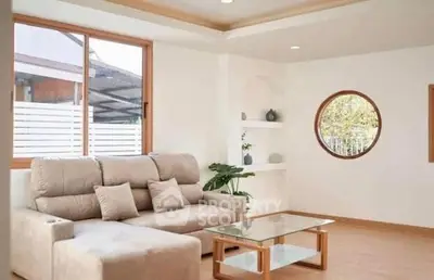 Modern living room with beige sofa and glass coffee table, featuring large windows and stylish decor.