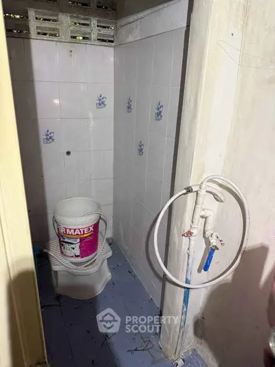 Compact bathroom with basic fixtures and tiled walls, featuring a bucket and hose setup.