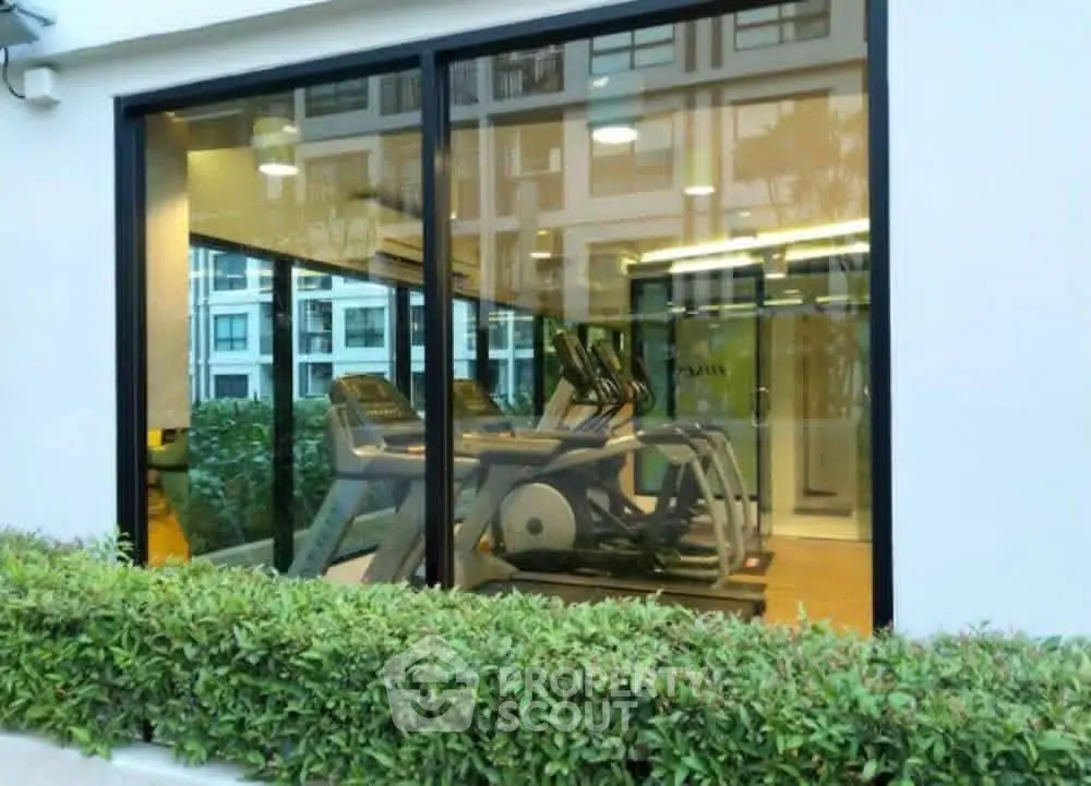 Modern gym with large windows in a residential building, showcasing fitness equipment and lush greenery outside.